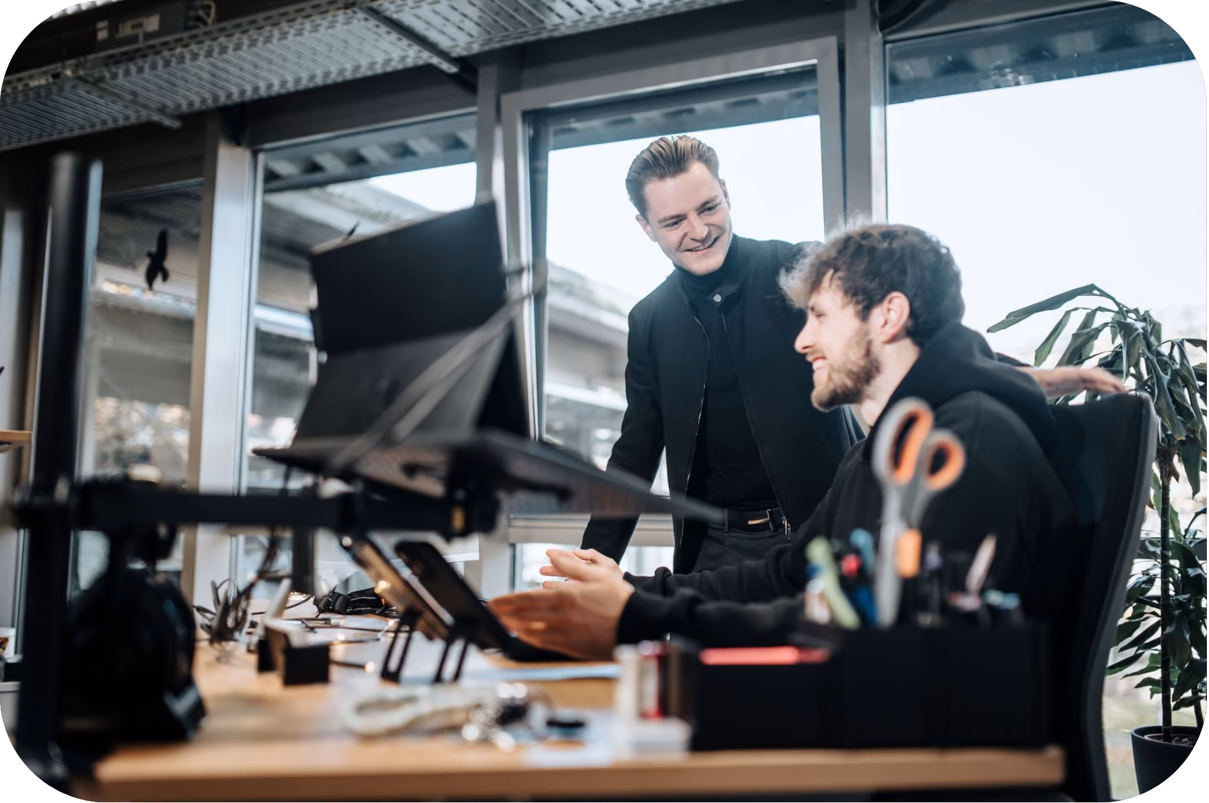 Two men in a modern office setting, one sitting and gesturing as he talks while the other stands behind him smiling, with a large drone on the desk in front of them.