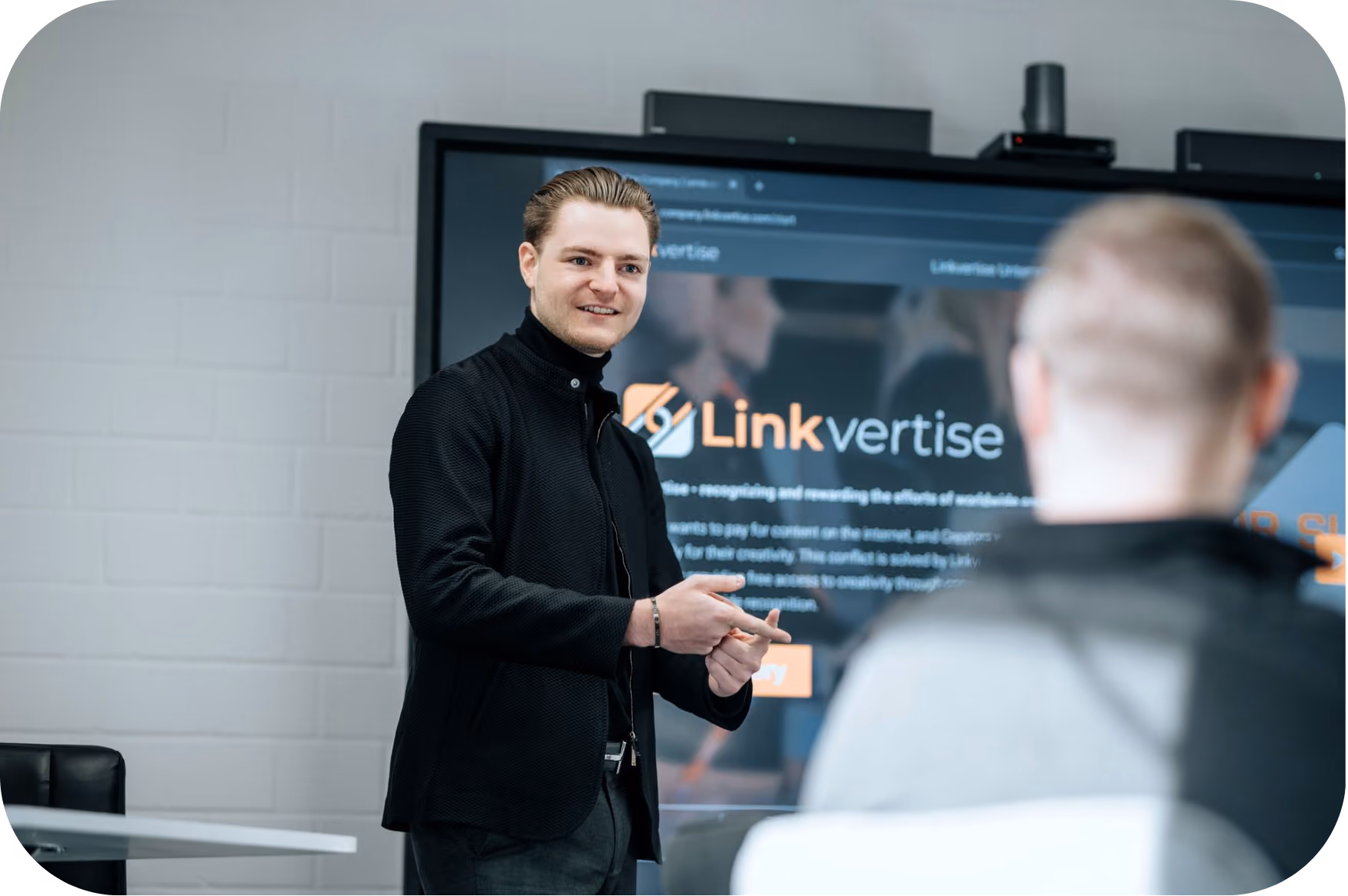 Young man in a black jacket standing and gesturing during a presentation with a large screen showing the Linkvertise logo and text behind him, while another person with a blurred head is seated facing him.