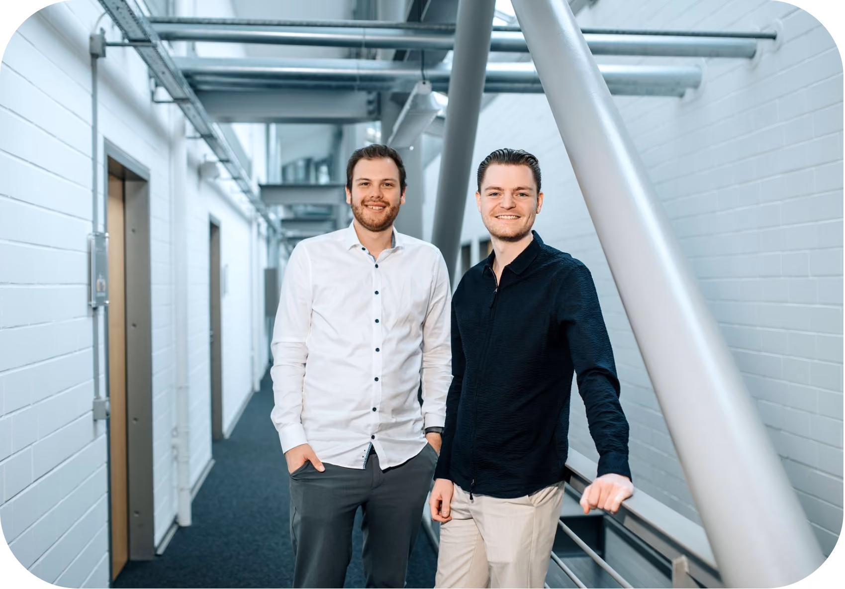 Two young men standing in a modern hallway with white walls and exposed pipes; the man on the left wears a white button-up shirt and gray pants, and the man on the right wears a black button-up shirt and light-colored pants, smiling at the camera.