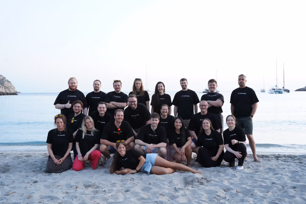 Group of twenty people posing on a sandy beach by the sea, wearing black t-shirts and smiling, with boats visible in the water in the background.